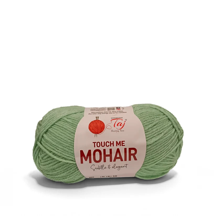 A skein of sage green mohair yarn with a cream-colored paper label that reads 'TOUCH ME MOHAIR'